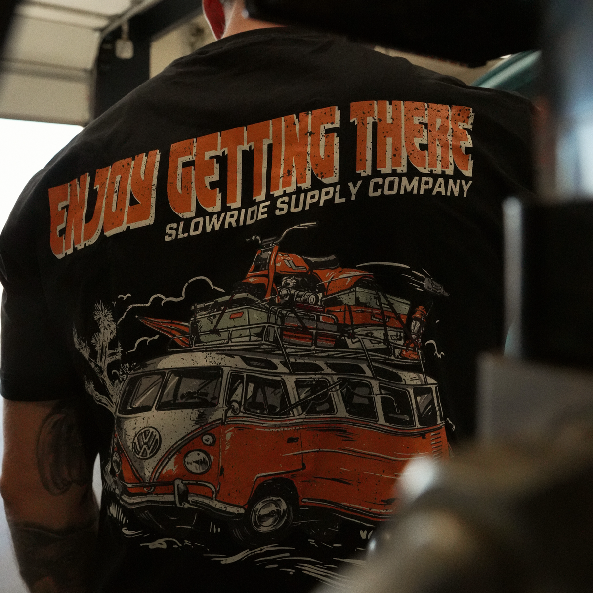 Person wearing Road Trip Black Tee, Garage interior with concrete floor and overhead lighting, wearer shown from behind -