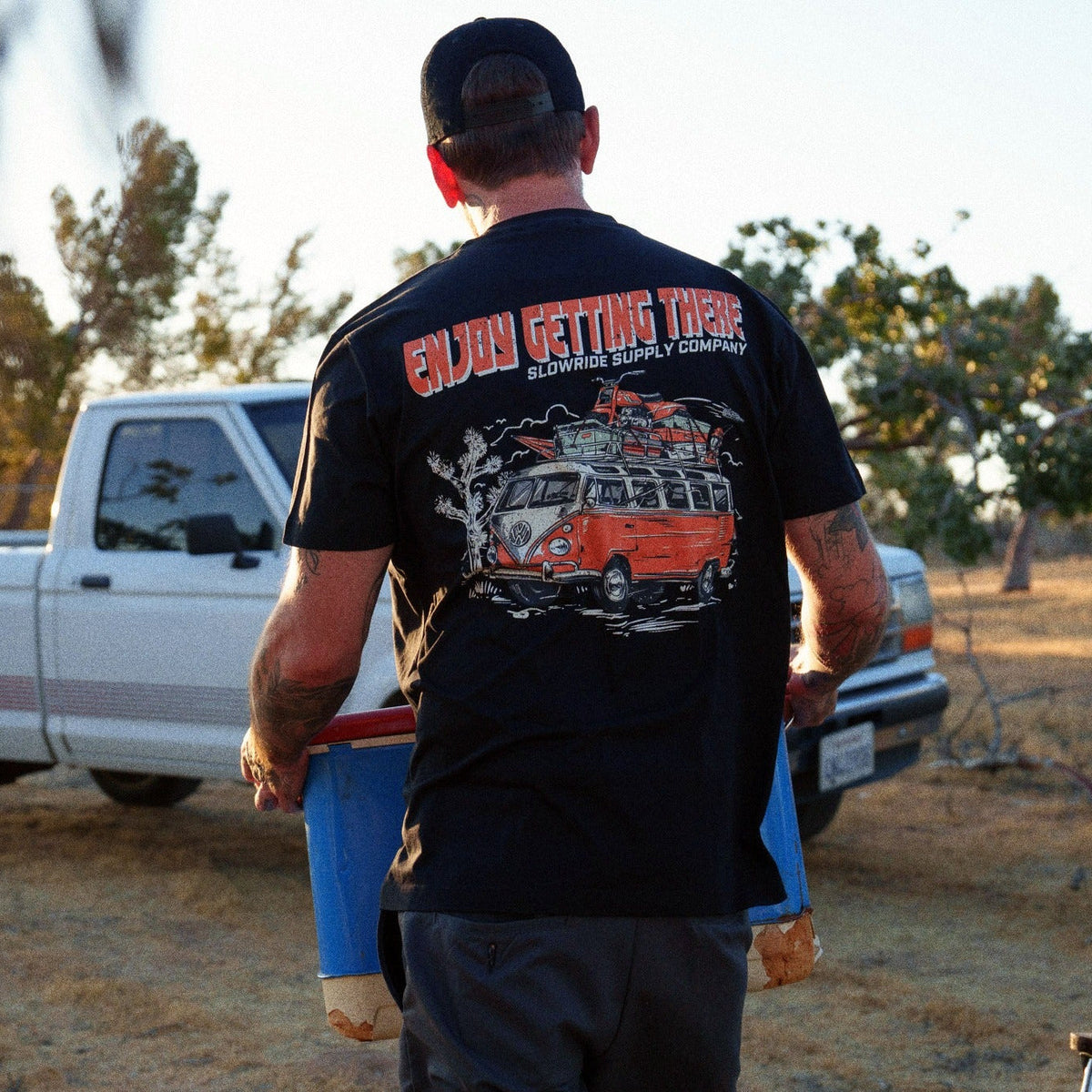 Person wearing Road Trip Black Tee, Desert landscape with parked vehicles and scattered trees - Slowride Supply
