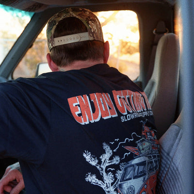 Person wearing Road Trip Black Tee, Inside a car on a desert highway, sunlit landscape visible through windshield - Slowride