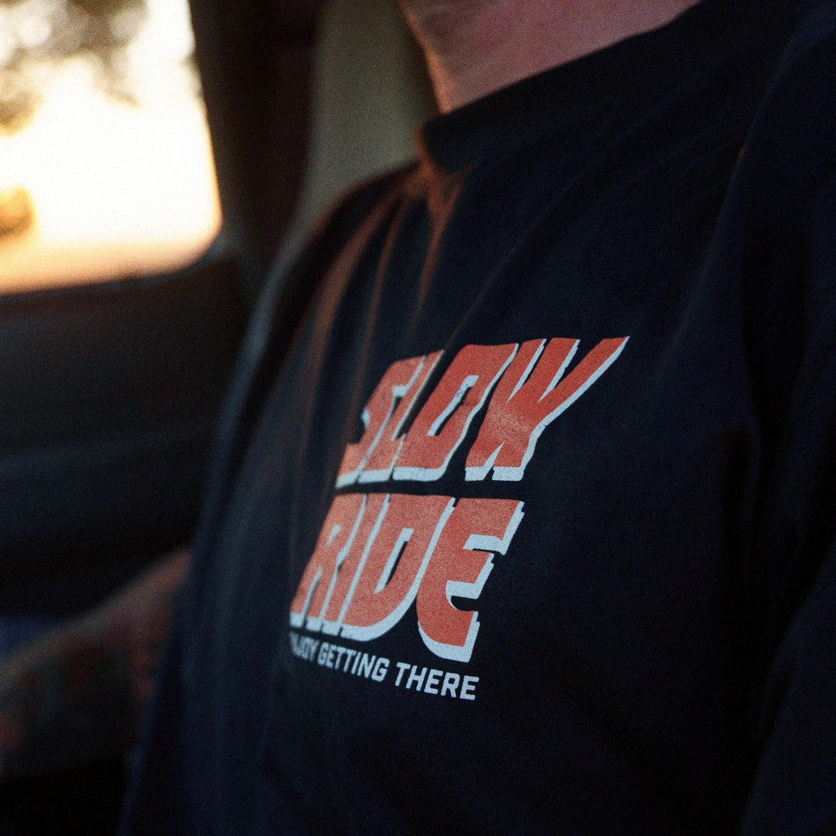 Person wearing Road Trip Black Tee, Person wearing black tee in warm garage or workshop setting with golden light - Slowride