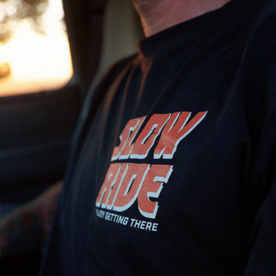 Person wearing Road Trip Black Tee, Person wearing black tee in warm garage or workshop setting with golden light - Slowride