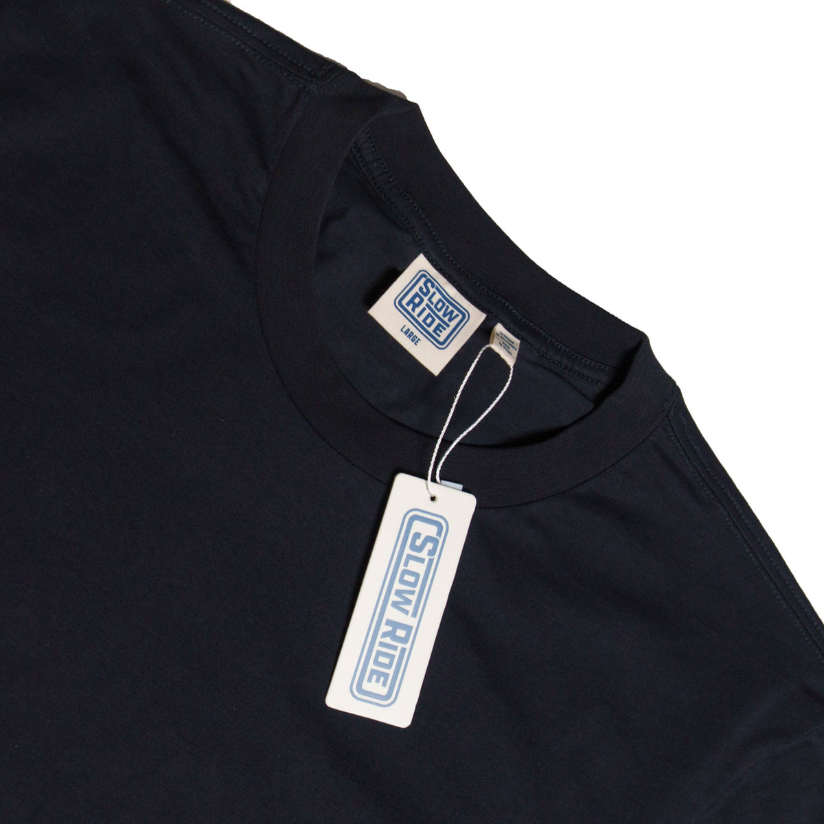 Care label on Road Trip Black Tee showing Cream-colored woven label with blue border and text on neckline, matching hang tag